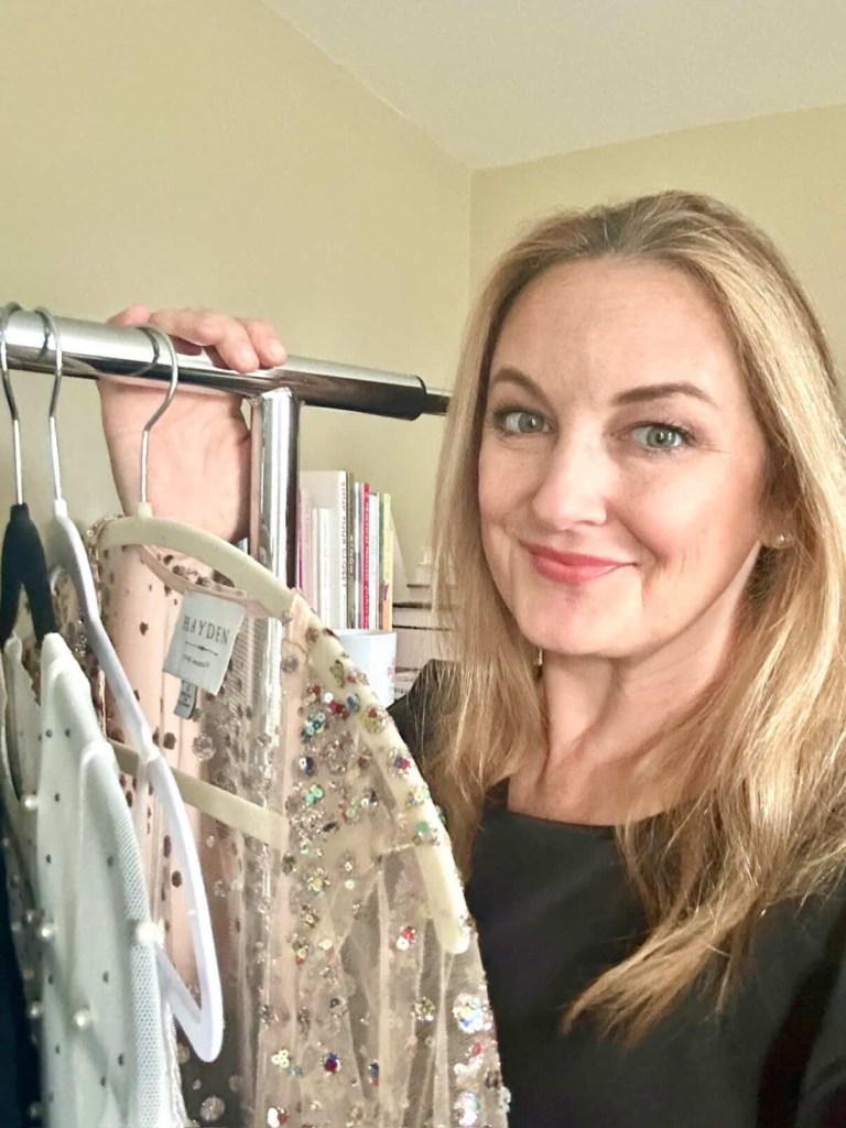 A woman holding a clothing rack with embellished dresses, smiling at the camera in a well-lit room.